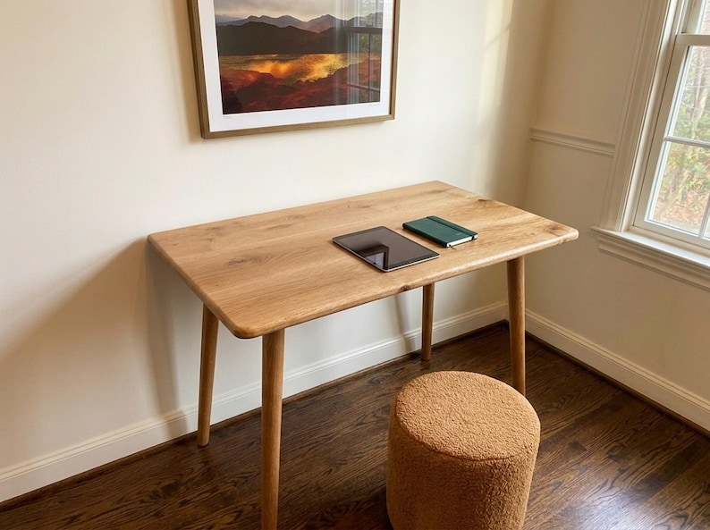 May include: A light-colored wooden desk with rounded corners and four tapered legs. A black tablet and a green notebook rest on the desk. A small, round, tan-colored stool sits below the desk.