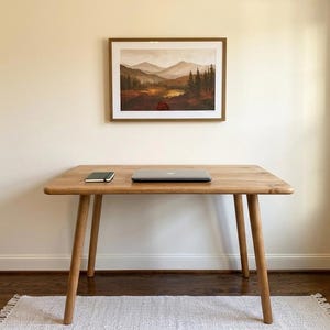 May include: A wooden desk with a laptop and notebook on top, beneath a framed landscape painting. The desk has four angled legs and a light wood finish. The painting depicts a mountain scene with trees and a lake.