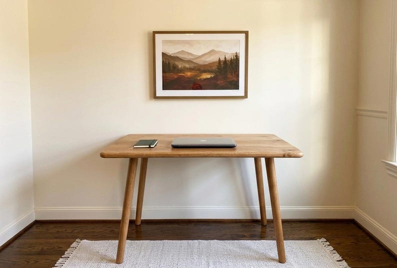 May include: A wooden desk with a laptop and notebook on top, beneath a framed landscape painting. The desk has four angled legs and a light wood finish. The painting depicts a mountain scene with trees and a lake.