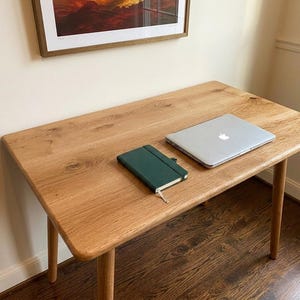 May include: A light-colored wooden desk with rounded corners and four legs. A closed silver laptop and a dark green notebook are on the desk. The desk is set against a beige wall with a framed picture.