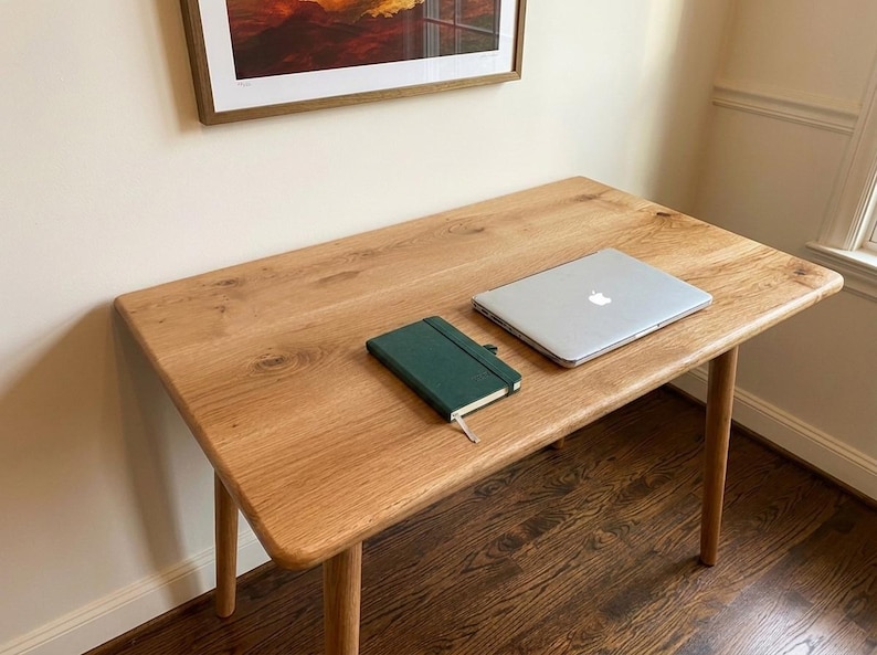 May include: A light-colored wooden desk with rounded corners and four legs. A closed silver laptop and a dark green notebook are on the desk. The desk is set against a beige wall with a framed picture.