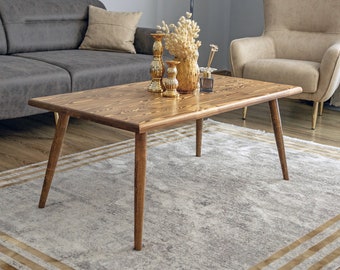 May include: A wooden coffee table with a rectangular top and four legs. The table is sitting on a rug with a light gray background and brown stripes. There are gold decorative items on the table, including a vase with dried flowers and candlesticks.