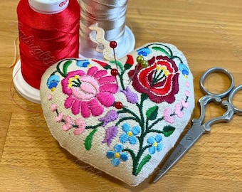 May include: A heart-shaped pincushion with embroidered flowers in shades of pink, red, blue, and green. The pincushion is filled with pins and is sitting on a wooden surface next to two spools of thread and a pair of scissors.