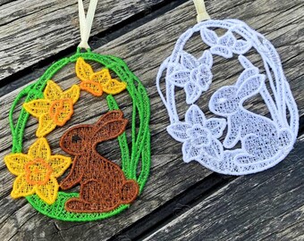 May include: Two Easter egg ornaments with a bunny and floral design. One egg is green with a brown bunny and yellow flowers. The other is white with a white bunny and white flowers. Both have a cream-colored ribbon for hanging.