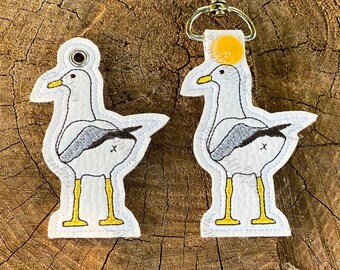 May include: Two white seagull keychains with black and gray markings. The keychains are made of felt and have a white background. One keychain has a yellow button on the top.