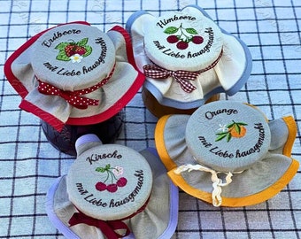 May include: Four glass jars with fabric lids, each with a different embroidered design and text. The lids are decorated with red, yellow, blue, and white ribbon. The jars are labeled "Erdbeere mit Liebe hausgemacht", "Himbeere mit Liebe hausgemacht", "Kirsche mit Liebe hausgemacht", and "Orange mit Liebe hausgemacht".