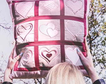 Op de afbeelding: Een vierkant sierkussen met een raster van negen panelen, elk met een uniek geborduurd hartontwerp in tinten roze en donkerrood. De stof is lichtroze met donkerrode randen die de afzonderlijke vierkanten met hartmotieven scheiden.