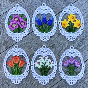 May include: Six white lace oval ornaments, each with a different embroidered flower arrangement. The flowers include purple hellebores, blue grape hyacinths, yellow daffodils, orange tulips, white lilies, and purple crocuses. The ornaments have a delicate, openwork design.