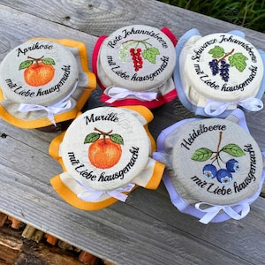 Puede incluir: Una colección de tapas de tarros bordadas con ilustraciones de frutas y texto en alemán. Cada tapa presenta una fruta diferente: albaricoque, grosella roja, grosella negra y arándano. Las tapas están adornadas con varios colores y atadas con cintas blancas.