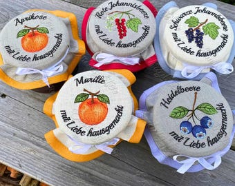 May include: A collection of embroidered jar covers with fruit illustrations and German text. Each cover features a different fruit: apricot, red currant, black currant, apricot, and blueberry. The covers are trimmed with various colors and tied with white ribbons.