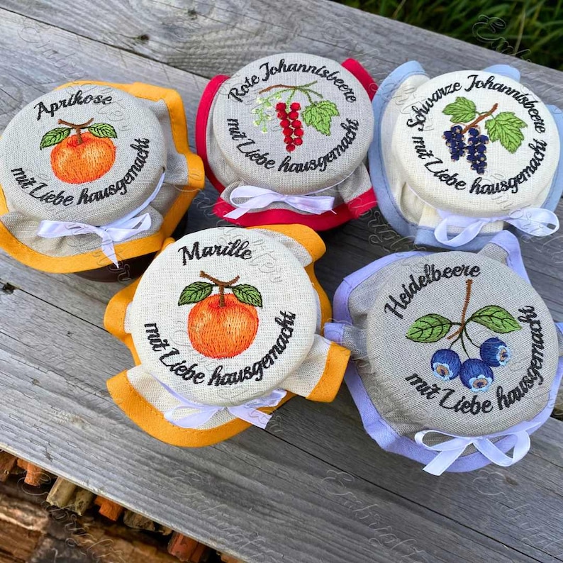 May include: A collection of embroidered jar covers with fruit illustrations and German text. Each cover features a different fruit: apricot, red currant, black currant, apricot, and blueberry. The covers are trimmed with various colors and tied with white ribbons.