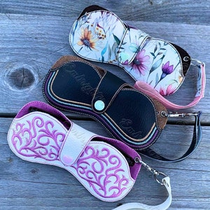 May include: Three eyeglass cases with wrist straps. The top case features a white background with a colorful floral print. The middle case is black with multi-colored stitching and a brown accent. The bottom case is white with pink embroidery.