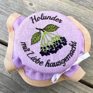 May include: A purple fabric jar lid with a brown trim and a white ribbon. The lid is embroidered with the text "Holunder mit Liebe hausgemacht" and a green sprig of elderberry with black berries.