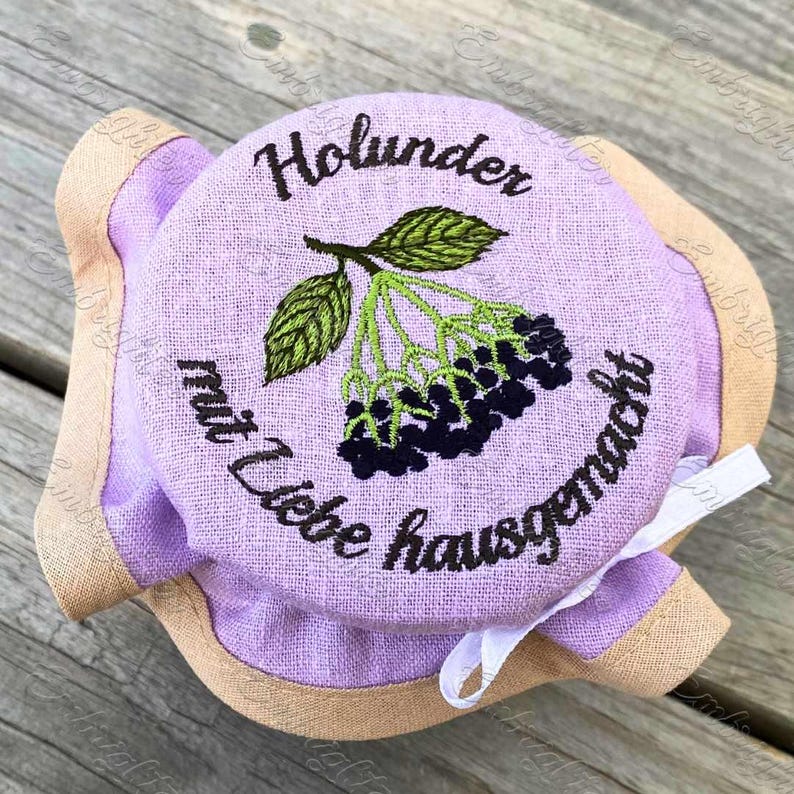 May include: A purple fabric jar lid with a brown trim and a white ribbon. The lid is embroidered with the text "Holunder mit Liebe hausgemacht" and a green sprig of elderberry with black berries.