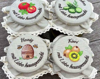 May include: Four jar lids with embroidered designs. Each lid features a different image: tomatoes, cucumbers, a beehive with bees, and kiwis. The lids are gray with white lace trim and text in German.