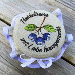 May include: Embroidered jar lid featuring a blueberry design with the text "Heidelbeere mit Liebe hausgemacht". The lid is surrounded by a light purple fabric and tied with a white ribbon.