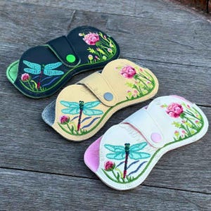 Può includere: Tre astucci ricamati per occhiali in blu navy, beige e bianco. Ogni astuccio presenta un design di libellula e floreale. Gli astucci hanno una chiusura a scatto e sono foderati con un materiale morbido. Gli astucci sono su una superficie di legno.