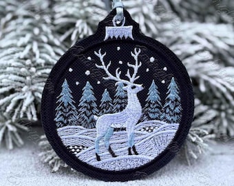 May include: Embroidered Christmas ornament with a black background featuring a winter scene. A white deer stands in a snowy landscape with pine trees. The ornament has a silver ribbon for hanging.