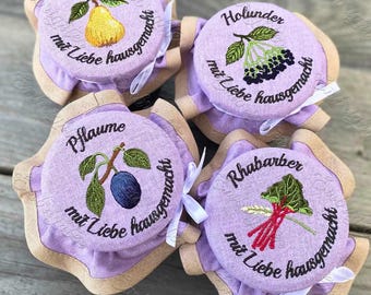 May include: Four purple fabric jars with embroidered lids. The lids have different fruit designs and the text "mit Liebe hausgemacht" which translates to "made with love homemade". The jars are labeled "Birne", "Pflaume", "Holunder", and "Rhabarber".