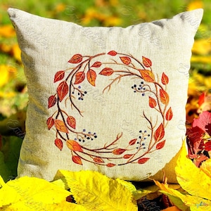 May include: A beige cushion featuring an embroidered autumnal wreath design. The wreath is made of brown branches with orange and red leaves and blue berries.