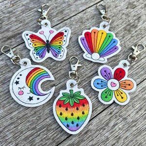 May include: Five embroidered keychains featuring rainbow-coloured designs: a butterfly, a seashell with a pearl, a crescent moon with a face, a strawberry, and a flower. Each keychain is attached to a silver-toned metal lobster clasp.