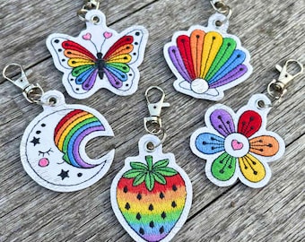 May include: Five embroidered keychains featuring rainbow-colored designs: a butterfly, a seashell with a pearl, a crescent moon with a face, a strawberry, and a flower. Each keychain is attached to a silver-toned metal lobster clasp.