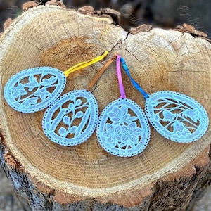 Könnte beinhalten: Vier blaue Spitzen-Ostereier-Anhänger mit Blumendesigns, die an bunten Bändern an einer Holzoberfläche hängen.