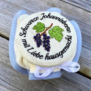 May include: A round, cream-colored jar lid with a blue trim and a white ribbon. The lid features an embroidered design of black currants, green leaves, and text in a circular pattern. The text reads "Schwarze Johannisbeere mit Liebe hausgemacht."