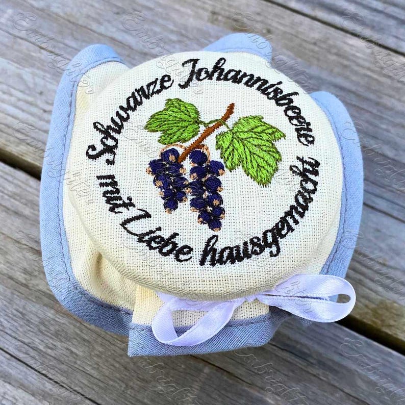 May include: A round, cream-colored jar lid with a blue trim and a white ribbon. The lid features an embroidered design of black currants, green leaves, and text in a circular pattern. The text reads "Schwarze Johannisbeere mit Liebe hausgemacht."