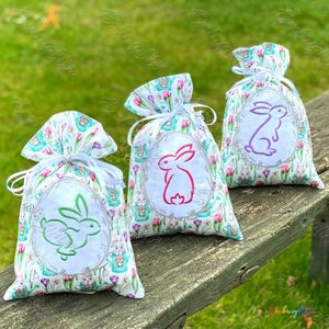 May include: Three white fabric Easter gift bags with a floral print and embroidered bunnies in pink, green, and purple. The bags are tied with white ribbon and are sitting on a wooden surface.