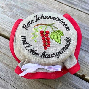 May include: A jar lid with embroidered text and a red currant design. The text reads "Rote Johannisbeere mit Liebe hausgemacht". The lid is beige with a red trim and a white ribbon.