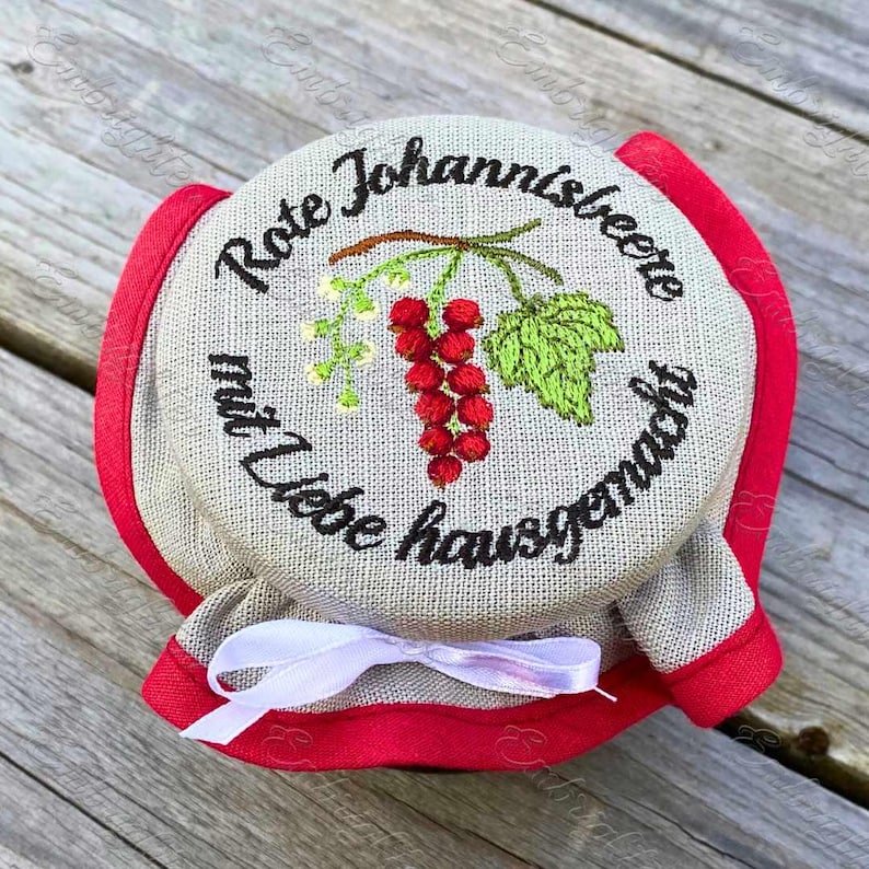 May include: A jar lid with embroidered text and a red currant design. The text reads "Rote Johannisbeere mit Liebe hausgemacht". The lid is beige with a red trim and a white ribbon.