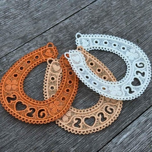 Puede incluir: Tres adornos de encaje en forma de herradura en tonos naranja, beige y blanco. Cada herradura presenta el número "2026" y recortes en forma de corazón. El encaje tiene un diseño calado delicado con bordes decorativos.
