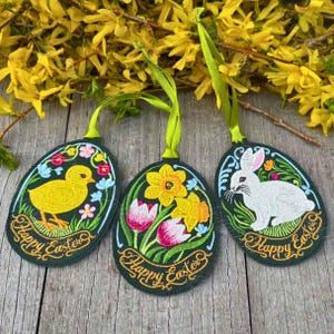 May include: Three embroidered Easter egg ornaments with green ribbon hangers. Each egg features a different design: a yellow chick, flowers, and a white bunny. The words "Happy Easter" are embroidered on each egg.