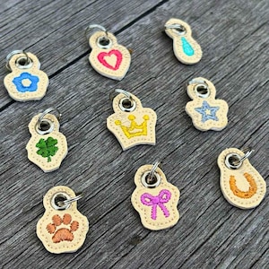 May include: A collection of small, beige keychains with various embroidered designs, including a flower, heart, crown, star, four-leaf clover, paw print, bow, and horseshoe. Each keychain has a silver metal ring.