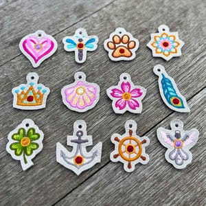 May include: A collection of embroidered patches in various shapes, including a heart, dragonfly, paw print, flower, crown, seashell, and feather. Each patch features detailed stitching and a small hole for attachment. The patches are in shades of pink, orange, blue, and green.