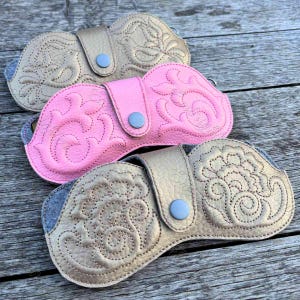 May include: Three eyeglass cases with snap closures, featuring embossed floral and bird patterns. The top case is beige, the middle case is bright pink, and the bottom case is metallic gold. Each case has a textured surface and a central snap button.