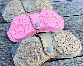 May include: Three eyeglass cases with snap closures, featuring embossed floral and bird patterns. The top case is beige, the middle case is bright pink, and the bottom case is metallic gold. Each case has a textured surface and a central snap button.