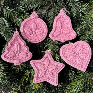 May include: Five dusty pink embroidered ornaments in the shapes of a round bauble, a bell, a Christmas tree, a star, and a heart. Each ornament features a stitched holly leaf and berry motif with intricate swirling patterns and a small ribbon loop.