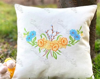 May include: A square, off-white pillow with an embroidered floral design. The embroidery features yellow daffodils, blue flowers, and green leaves. The pillow is placed on a bed of green grass.
