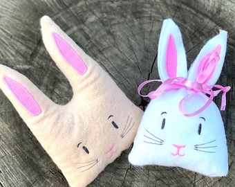 May include: Two plush bunny-shaped gift bags. One is beige with pink ears and the other is white with pink ears and a pink ribbon. Both have embroidered faces with black eyes and whiskers.