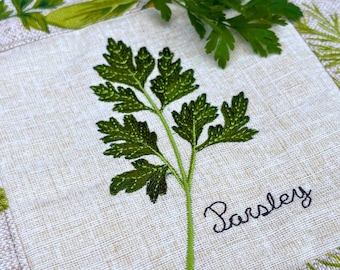 Può includere: Un quadrato di tessuto beige chiaro con un rametto di prezzemolo verde ricamato al centro. La parola Parsley è cucita in corsivo nero a destra del gambo. Il tessuto presenta una trama visibile su tutta la superficie.