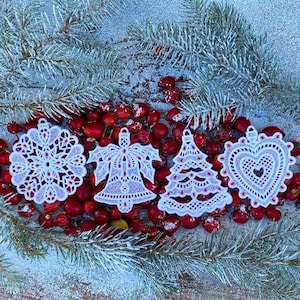Puede incluir: Cuatro adornos navideños de encaje blanco con detalles rojos. Los adornos son un copo de nieve, una campana, un árbol de Navidad y un corazón. Los adornos están sobre un fondo de bayas rojas y ramas de coníferas.