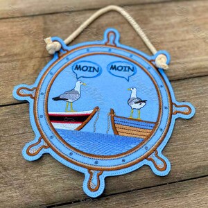 May include: A blue felt nautical wheel with two seagulls perched on boats. The seagulls are saying "Moin" in speech bubbles. The wheel has a brown rope hanging from the top.
