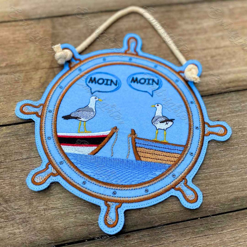 May include: A blue felt nautical wheel with two seagulls perched on boats. The seagulls are saying "Moin" in speech bubbles. The wheel has a brown rope hanging from the top.