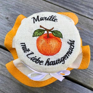 May include: A jar lid with an embroidered orange apricot and green leaves. The lid has the word "Marille" and the phrase "mit Liebe hausgemacht" in black script. A white ribbon is tied at the bottom, and the lid is surrounded by orange fabric.