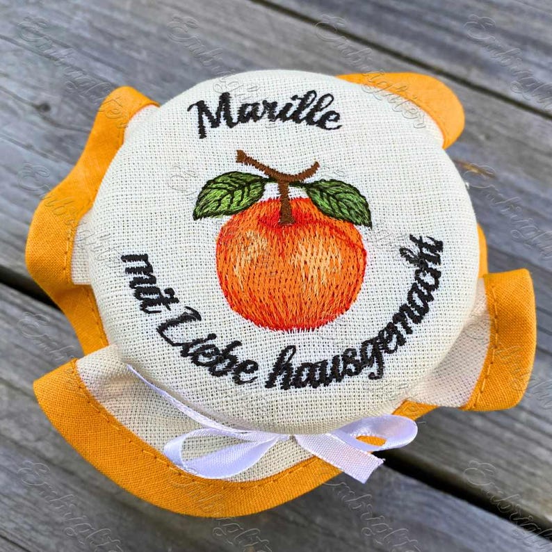 May include: A jar lid with an embroidered orange apricot and green leaves. The lid has the word "Marille" and the phrase "mit Liebe hausgemacht" in black script. A white ribbon is tied at the bottom, and the lid is surrounded by orange fabric.