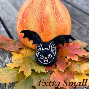May include: A black embroidered bat patch with white outlines, pointed ears, and small fangs, positioned in front of a large orange pumpkin and scattered autumn maple leaves. The text "Extra Small" is printed in black at the bottom of the image.