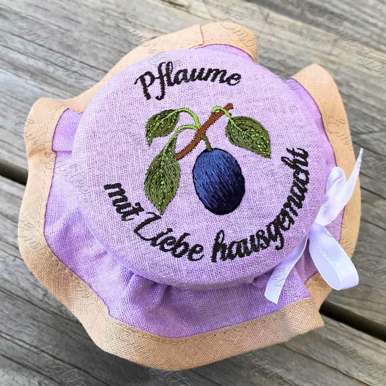 May include: A purple fabric jar lid with a brown embroidered plum and the text "Pflaume mit Liebe hausgemacht".