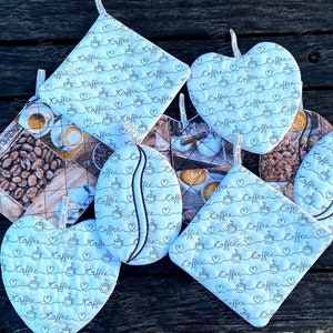 Puede incluir: Colección de agarraderas y manoplas acolchadas. Algunas tienen forma de corazón, otras cuadradas u ovaladas. La tela blanca está estampada con tazas de café, corazones y la palabra "Kaffee" en una línea continua. Algunas tienen imágenes de granos de café y bebidas.